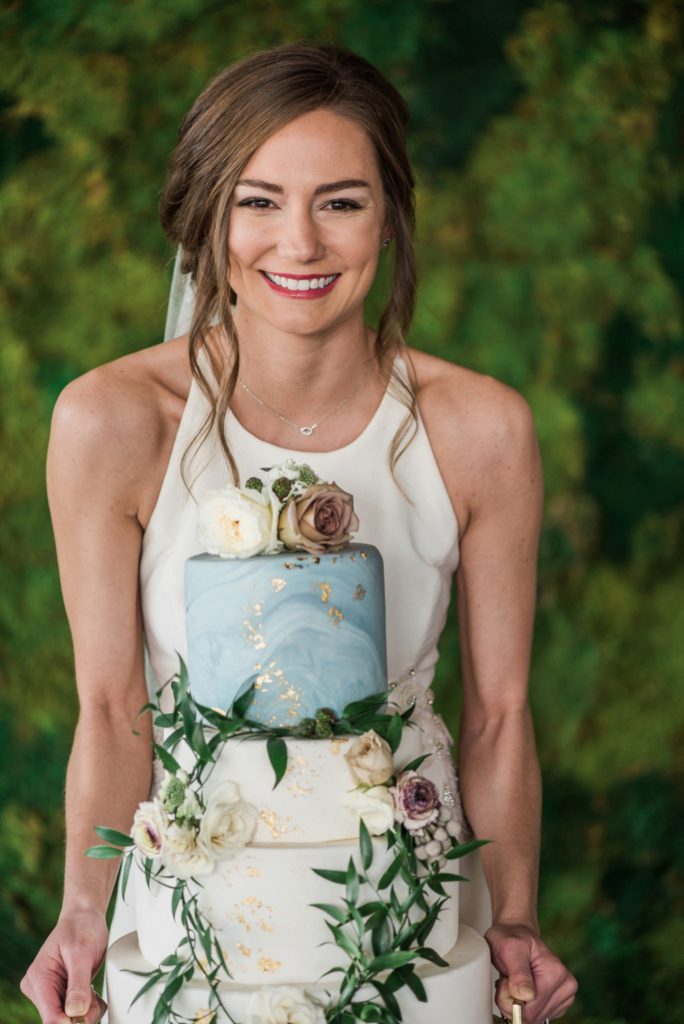 bride holding wedding cake and smiling with floral crown for cake at Moss | Denver
